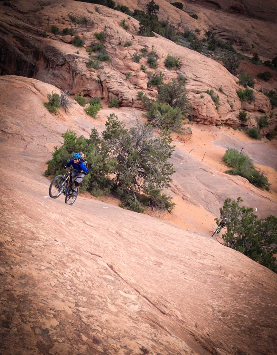 A mountain biker navigating a rocky, sloped terrain with shrubs and small trees in the background. The cyclist is wearing blue attire and a helmet, showcasing an adventurous outdoor setting. Slickrock mountain bike trail.