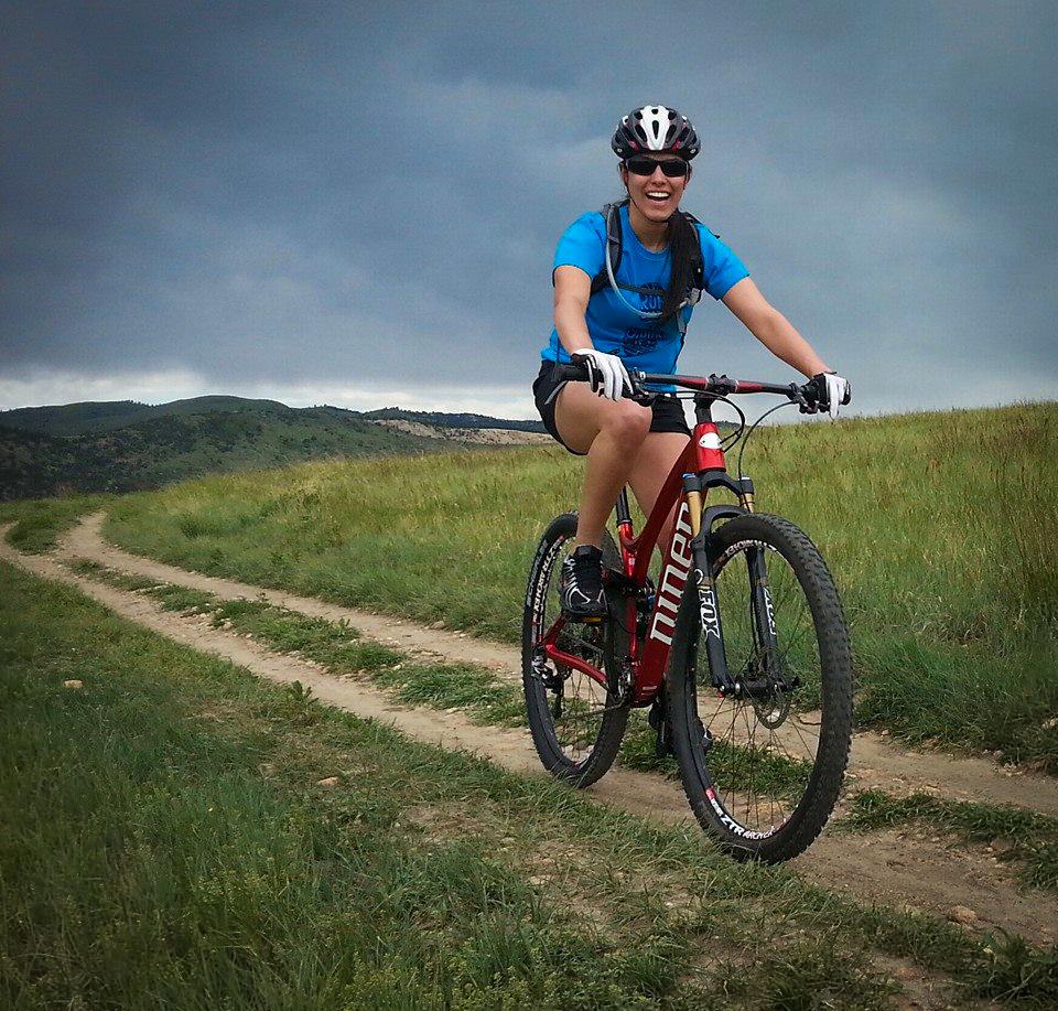 A woman riding a red mountain bike along a dirt path in a grassy landscape. She is wearing a blue shirt, black shorts, and protective eyewear. The sky is partly cloudy, creating a dramatic backdrop for her ride through the scenic terrain. Green Mountain mountain bike trail.
