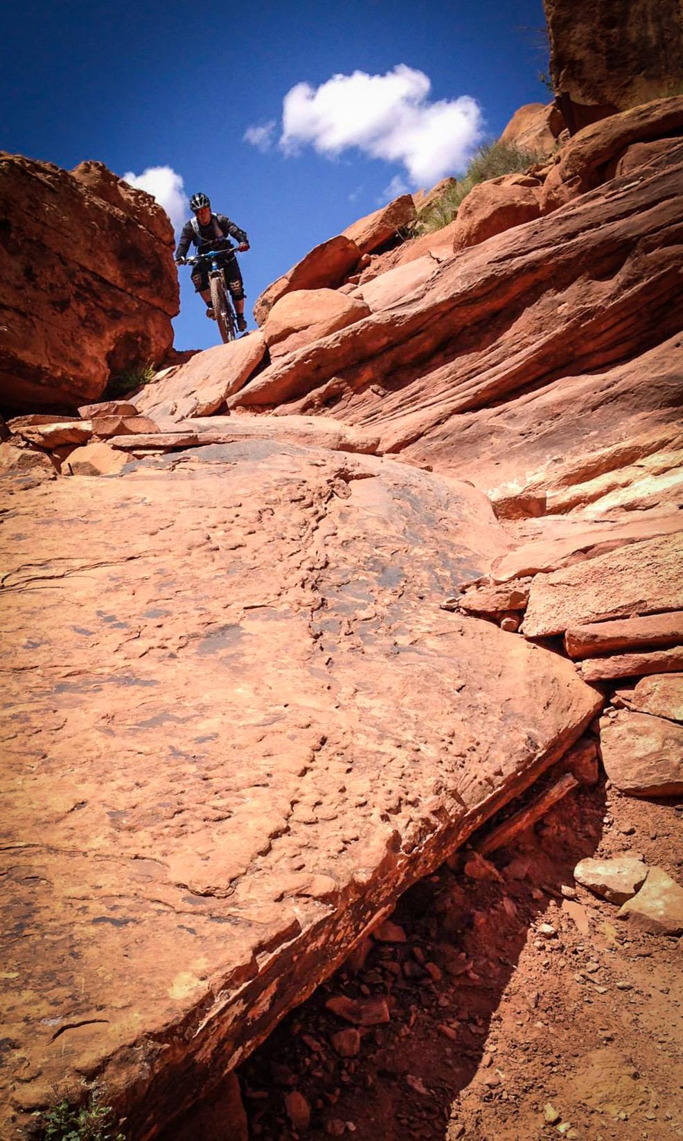 A mountain biker navigating a rocky trail surrounded by sunlit red rock formations and blue skies with scattered clouds. Porcupine Rim mountain bike trail.