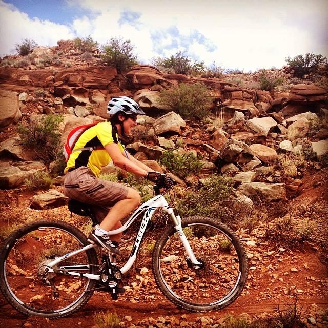 A person riding a mountain bike along a rocky trail, wearing a yellow and black jersey, shorts, and a helmet. The landscape features red rocks and sparse vegetation under a partly cloudy sky. Haystack Mountain mountain bike trail.