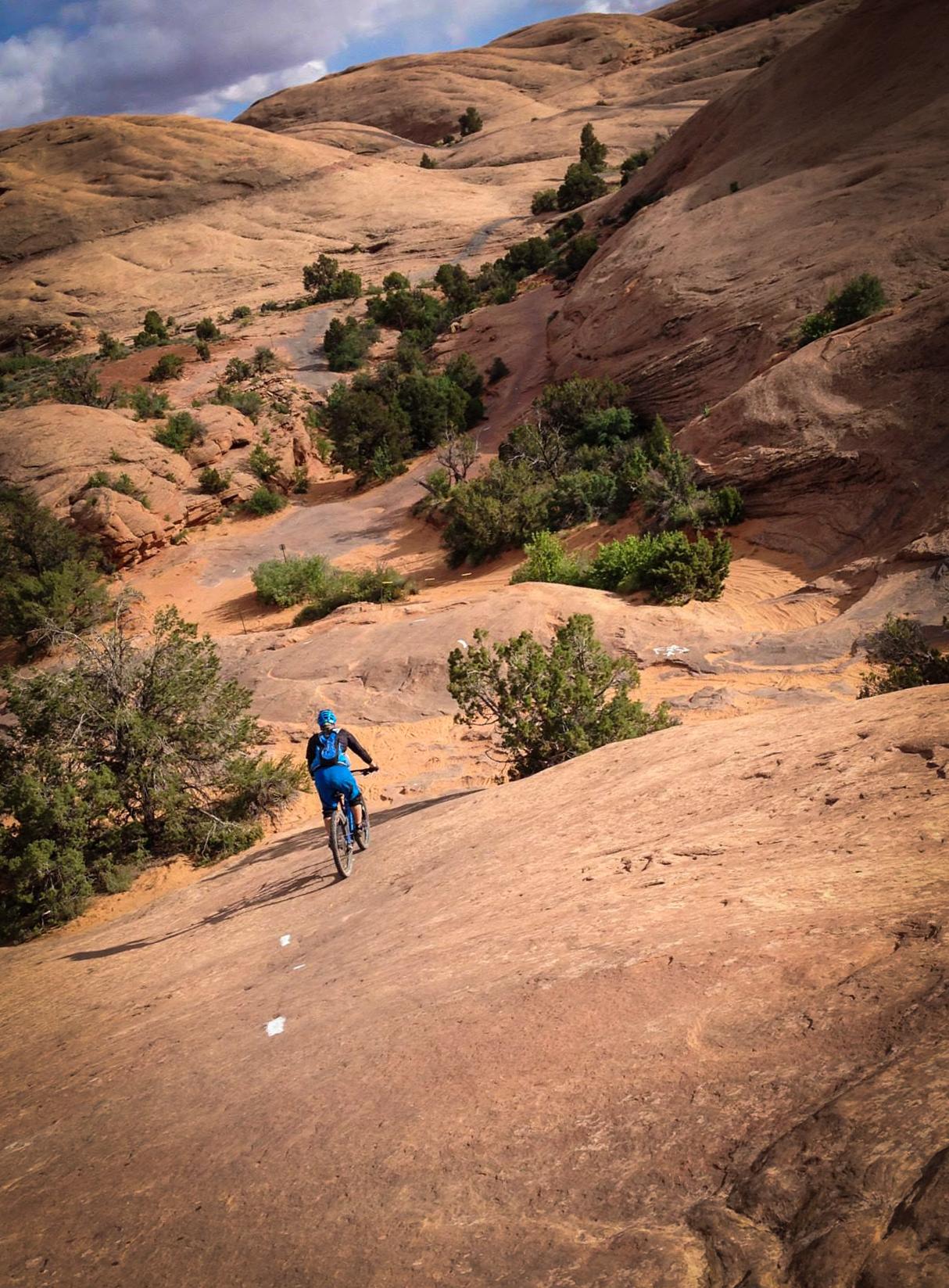 A mountain biker in a blue outfit rides down a rocky trail surrounded by rugged, sandy hills and sparse vegetation under a partly cloudy sky. Slickrock mountain bike trail.