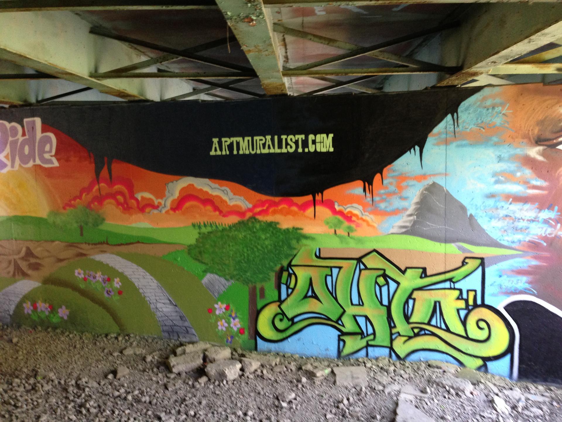 Colorful mural under a bridge featuring a scenic landscape with rolling hills, trees, and a mountain in the background. The artwork includes vibrant clouds and a path adorned with flowers. The mural also displays graffiti-style text, including the phrase "artmuralist.com" in bold lettering. Jermain Park mountain bike trail.