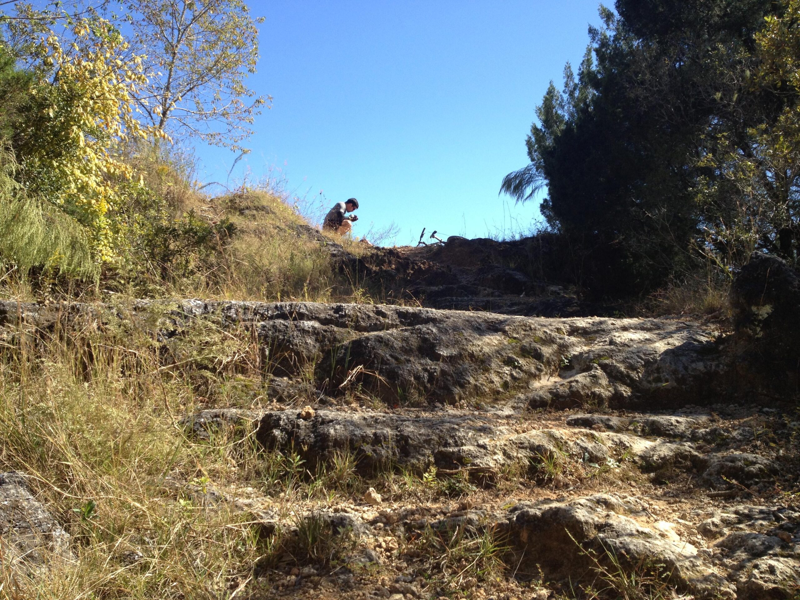 A person sitting on rocky terrain surrounded by grass and trees, with a clear blue sky above. Santos mountain bike trail.