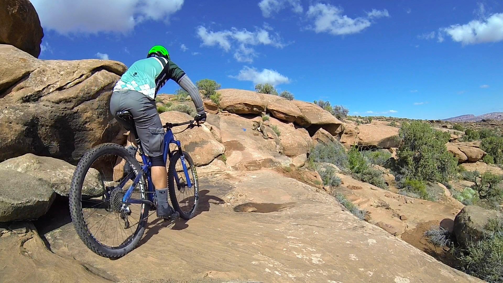 A mountain biker navigating rocky terrain with a blue bike and green helmet, surrounded by a natural landscape of shrubs and boulders under a clear blue sky with a few clouds. Captain Ahab mountain bike trail.