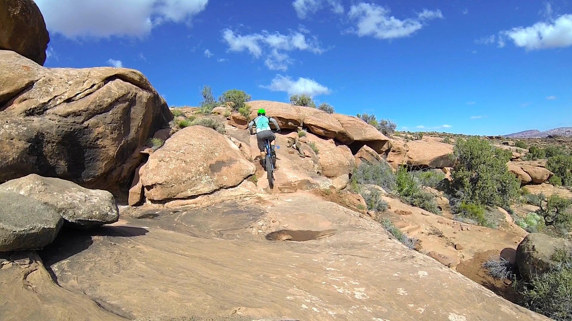 A mountain biker ascends a rocky trail surrounded by natural landscape, with large boulders and sparse vegetation under a bright blue sky with scattered clouds. Captain Ahab mountain bike trail.