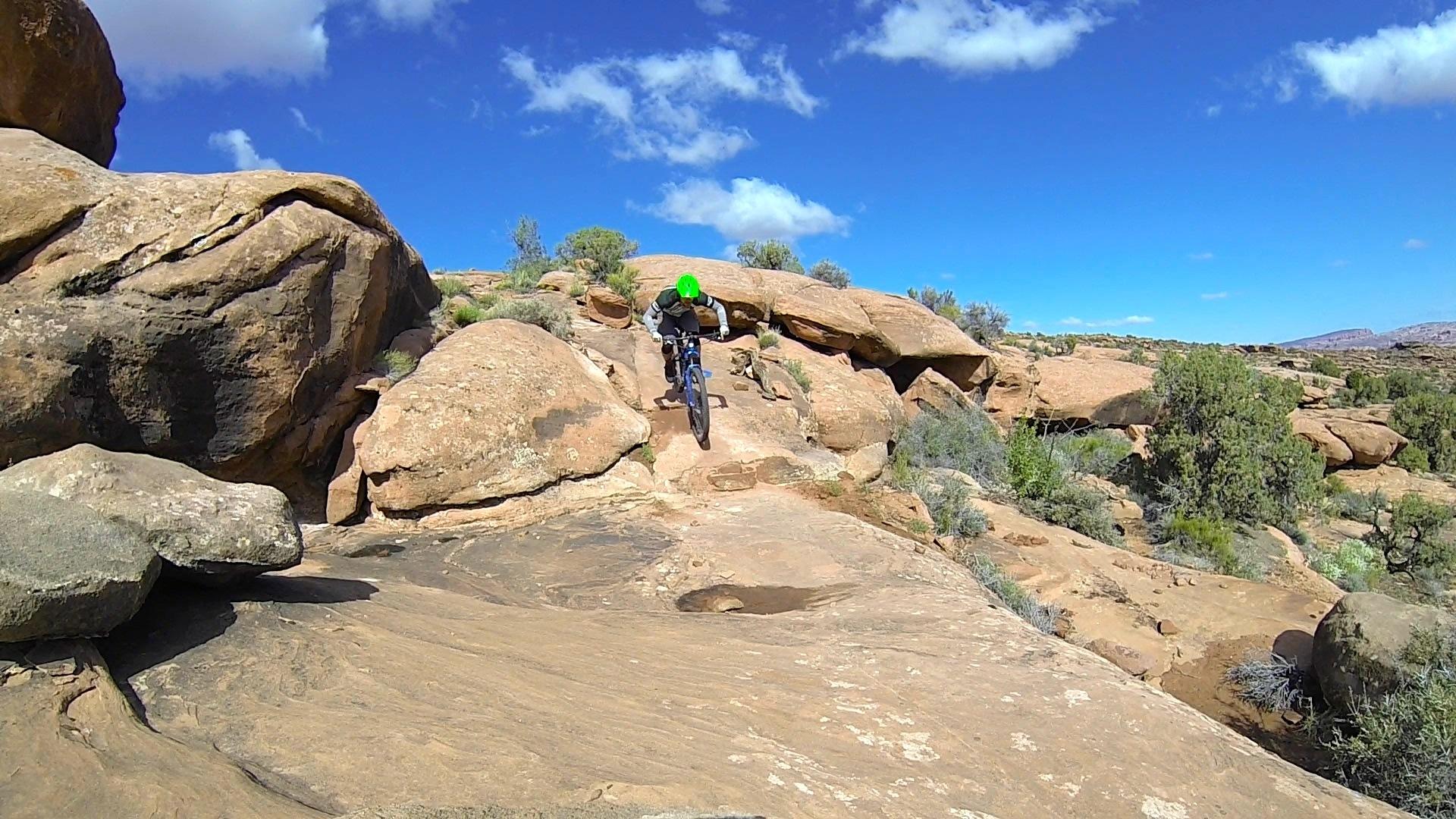 A mountain biker wearing a green helmet navigates a rocky terrain under a bright blue sky with scattered clouds. The rider is in mid-air as they descend a steep, rocky slope, surrounded by various shrubs and large boulders. Captain Ahab mountain bike trail.