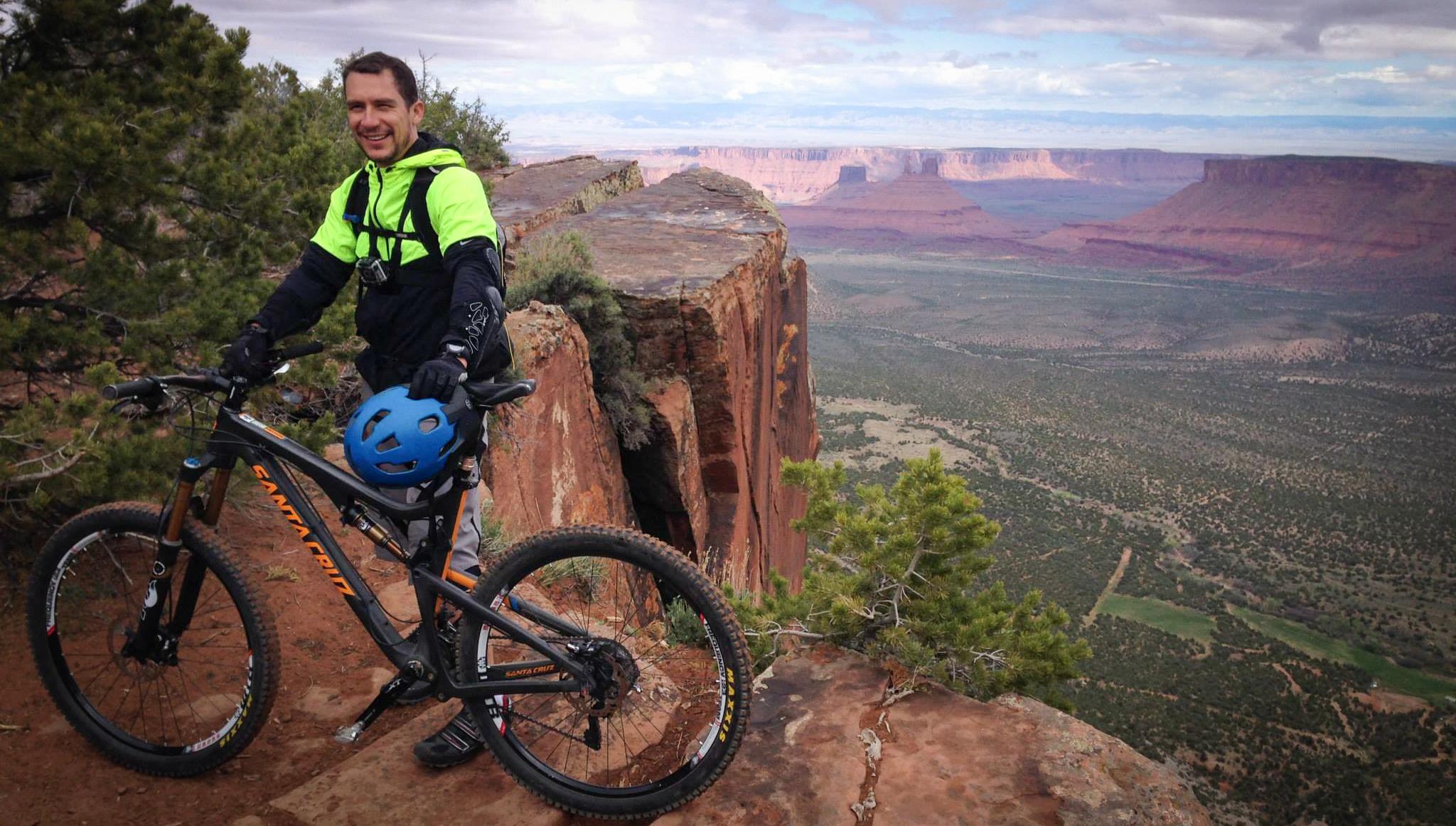 A mountain biker in a bright green jacket and blue helmet stands next to his bike on a rocky cliff, overlooking a vast valley with red rock formations and green vegetation in the distance. Porcupine Rim mountain bike trail.
