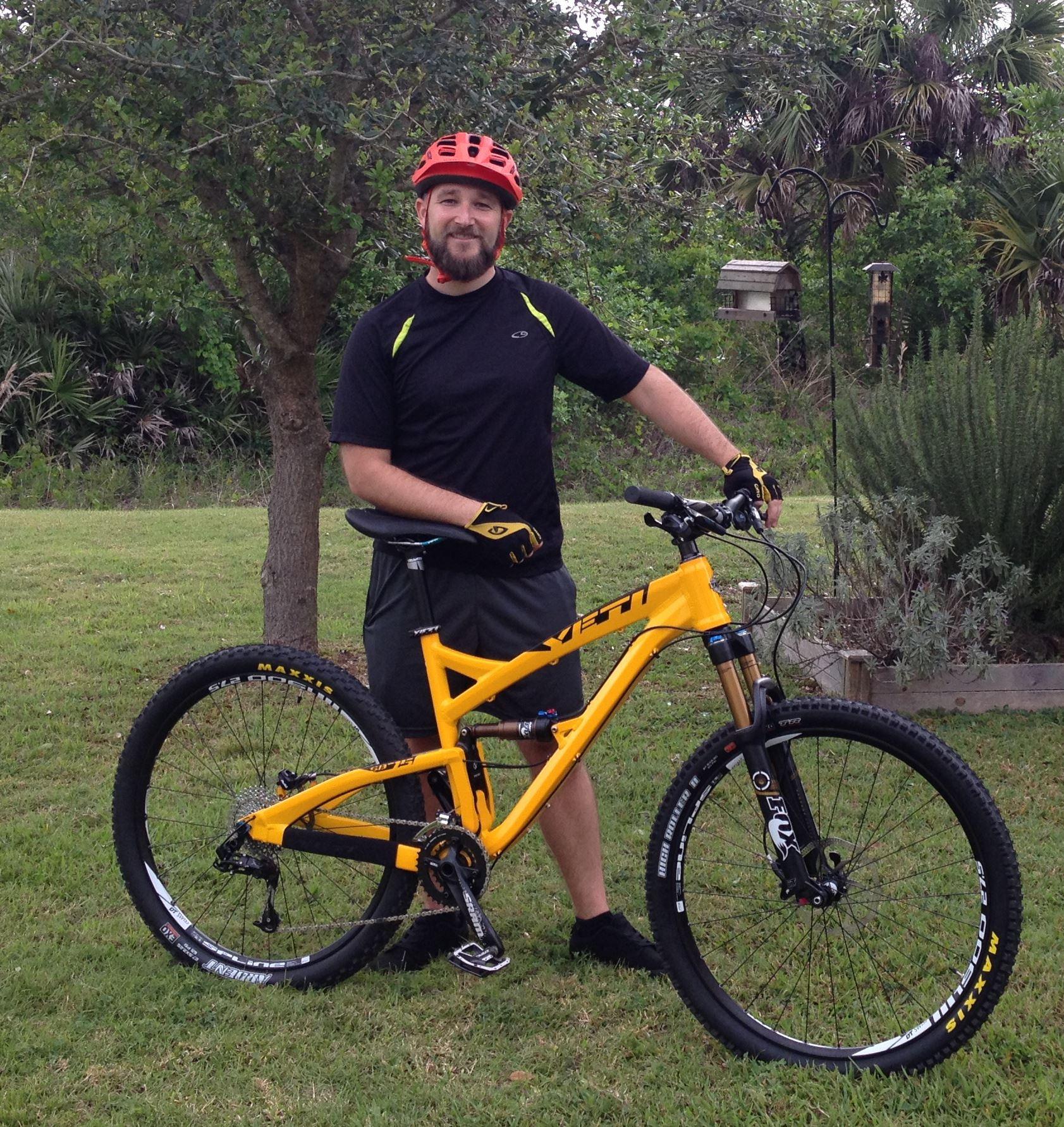 Yeti SB-75: A man stands next to a bright yellow mountain bike, wearing a red helmet, black shirt, and yellow gloves. He is positioned in a grassy area with trees and shrubs in the background, smiling at the camera.