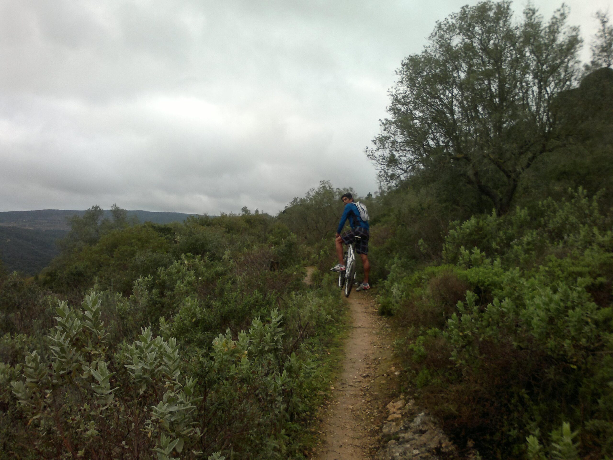 A person in a blue jacket is standing next to a white mountain bike on a dirt trail surrounded by lush greenery and low shrubs. The sky is overcast, and the landscape features rolling hills in the background. Arrabida mountain bike trail.