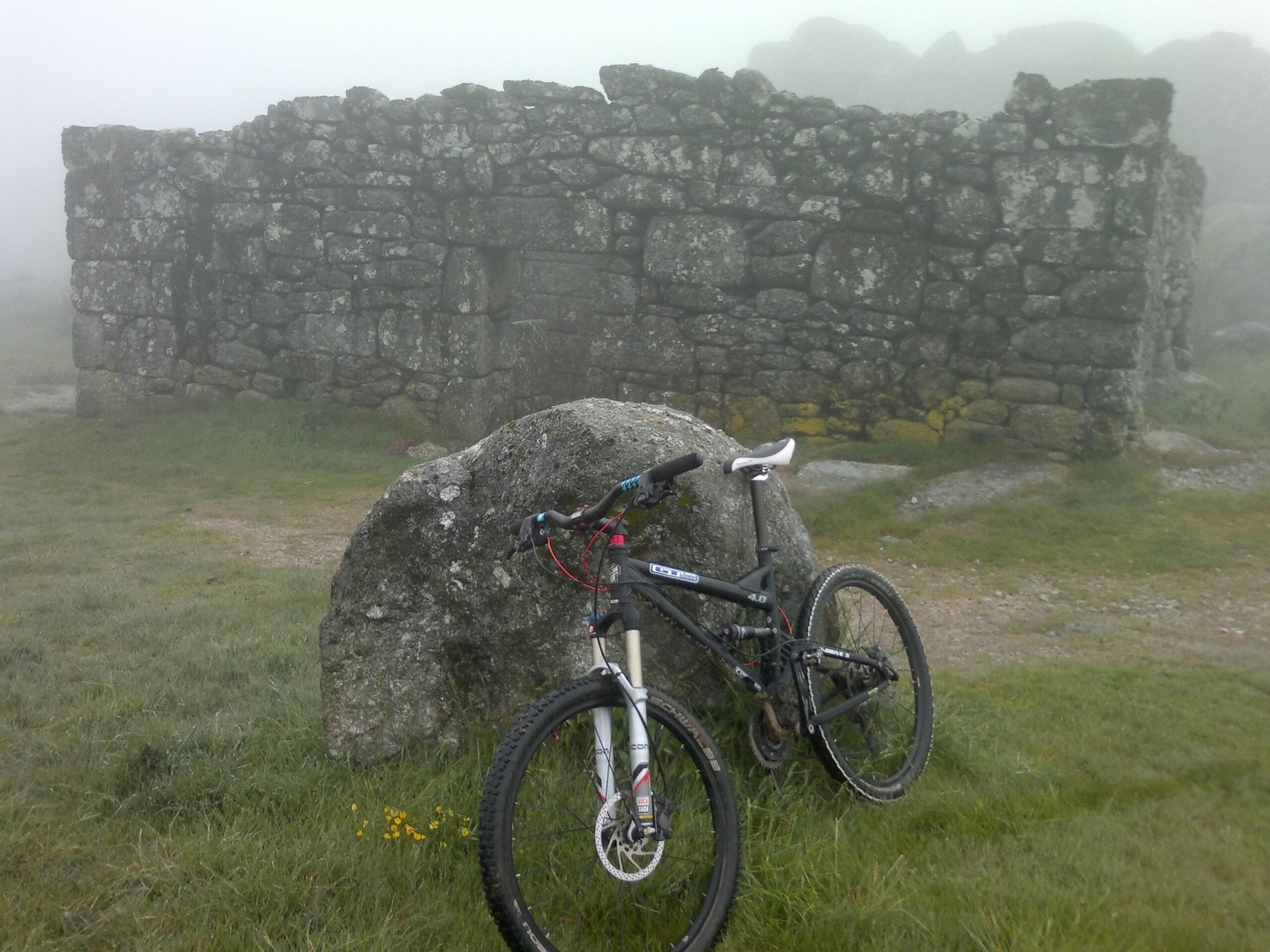 GT iDrive 5 4.0: A mountain bike leaning against a large rock, with a misty, fog-covered stone ruin in the background. The landscape features green grass and scattered stones, creating a serene outdoor atmosphere.