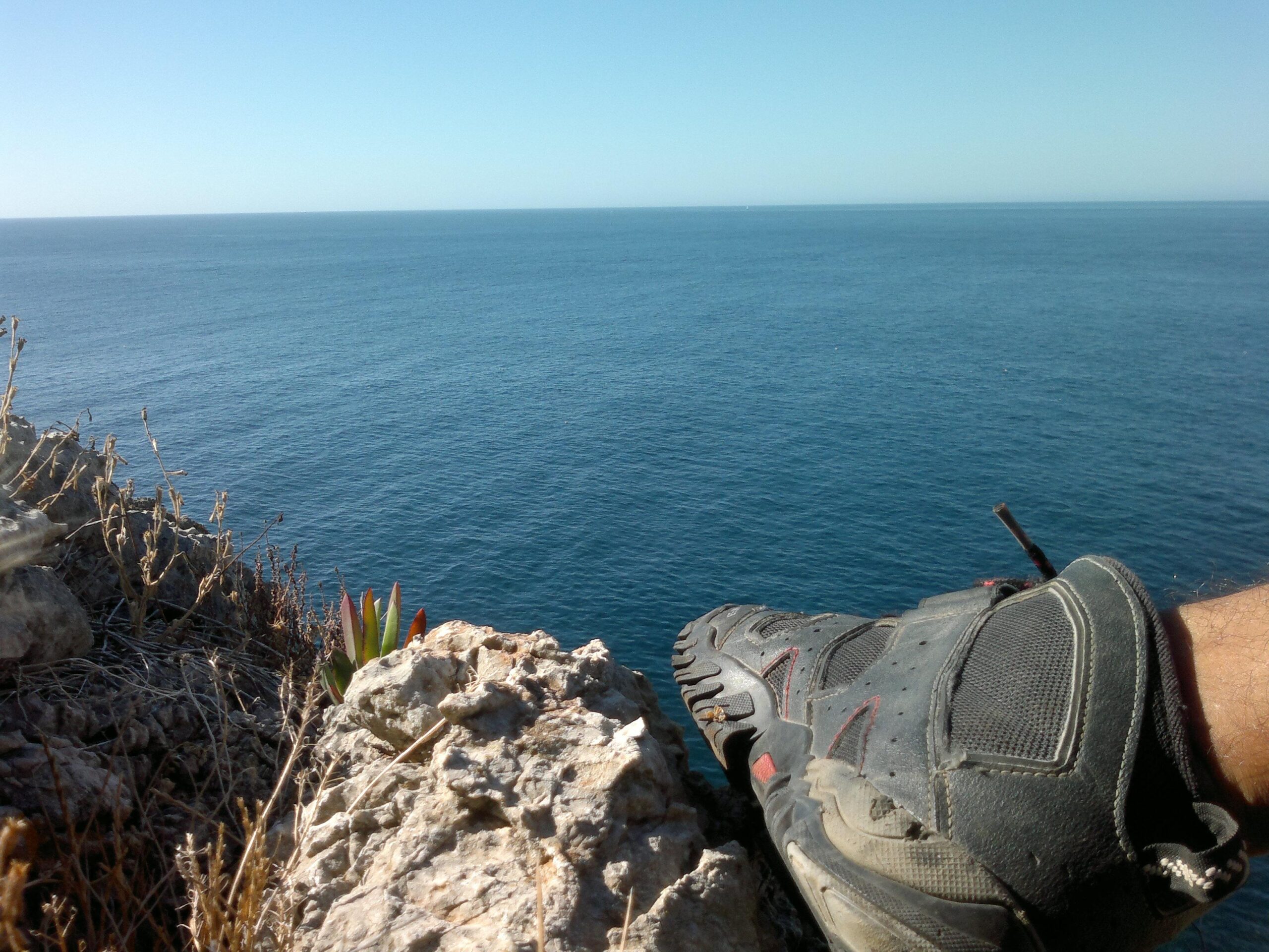 A close-up view of a hiker's shoe resting on a rocky ledge, overlooking a serene expanse of blue ocean under a clear sky. Small plants grow among the rocks, adding a touch of greenery to the scene. Arrabida mountain bike trail.
