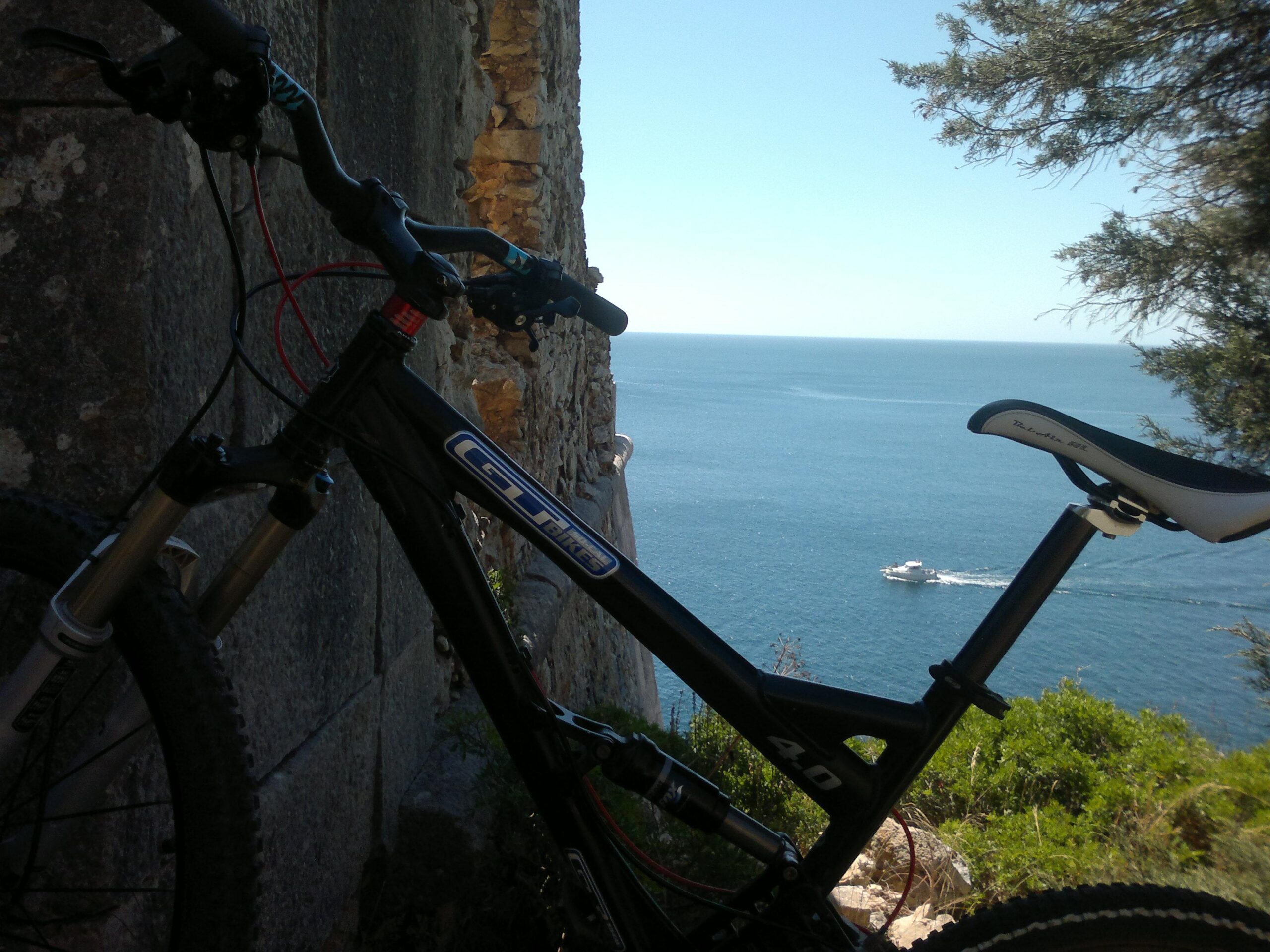 A mountain bike leaning against a stone wall with a view of the ocean and a boat in the distance, under a clear blue sky. Arrabida mountain bike trail.