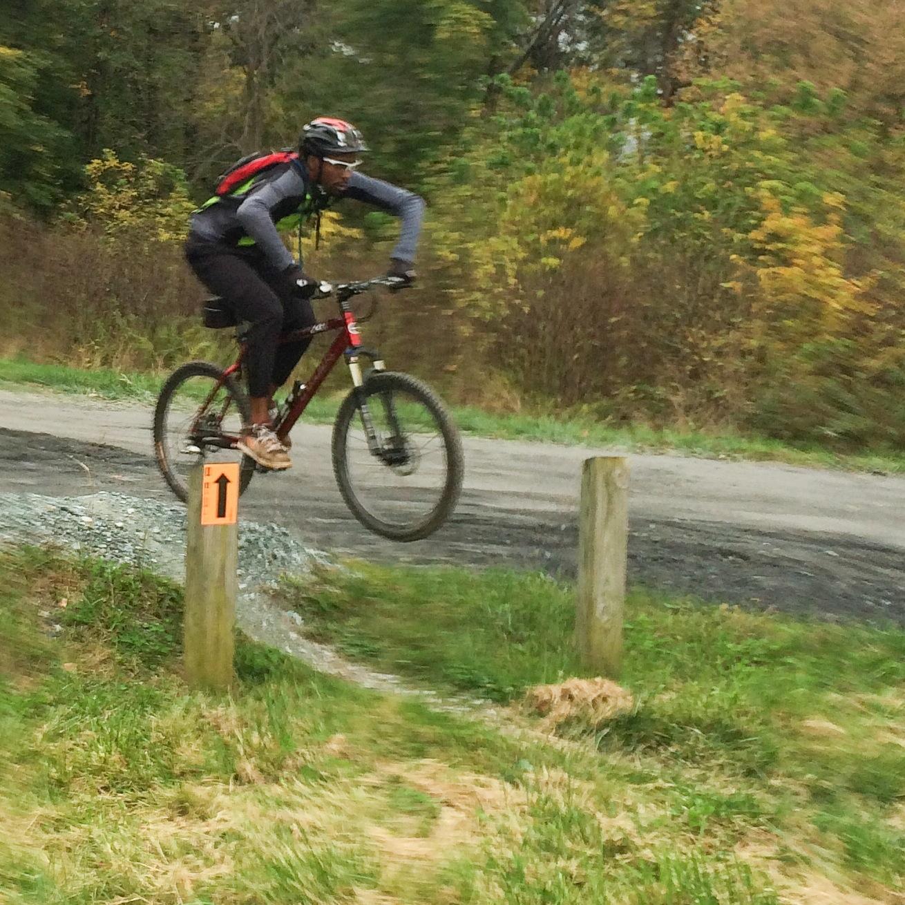 Trek 8000: A mountain biker in a helmet and protective gear is captured mid-jump over a small mound of dirt on a trail, with a marked post indicating the direction ahead. Surrounding foliage shows signs of autumn colors, creating a scenic outdoor environment.