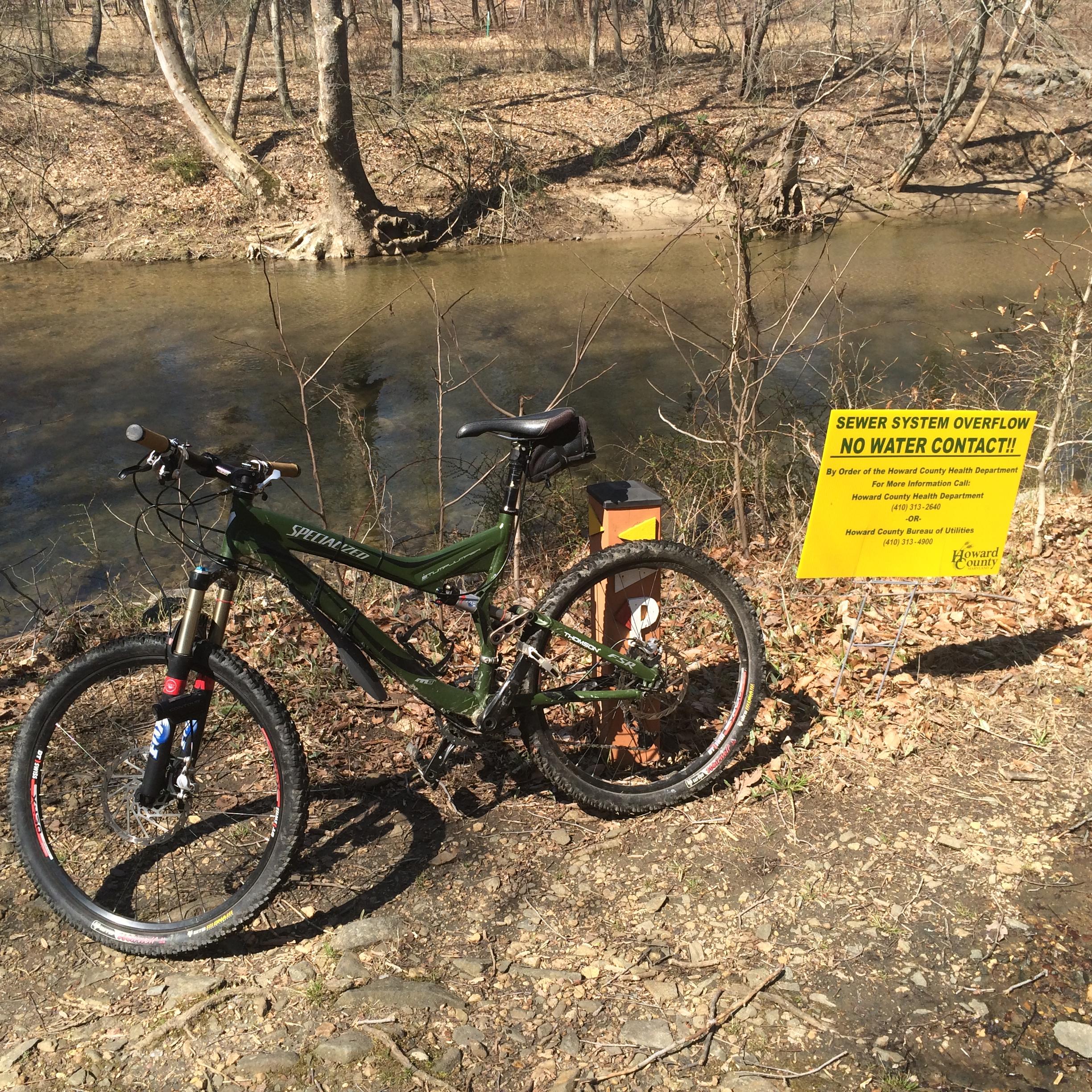Specialized Stumpjumper FSR: A mountain bike parked on a gravel path next to a clear creek. In the background, trees line the creek bank, and a bright yellow sign warns of a sewer system overflow, stating "NO WATER CONTACT!!" The area appears to be in a natural setting with dry leaves scattered on the ground.