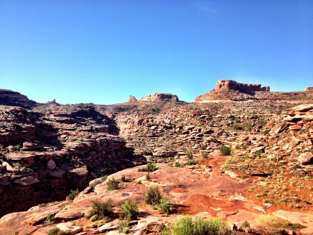 A wide view of a rocky landscape under a clear blue sky, featuring layered rock formations and sparse vegetation, suggesting a rugged terrain typical of desert environments. Hymasa mountain bike trail.