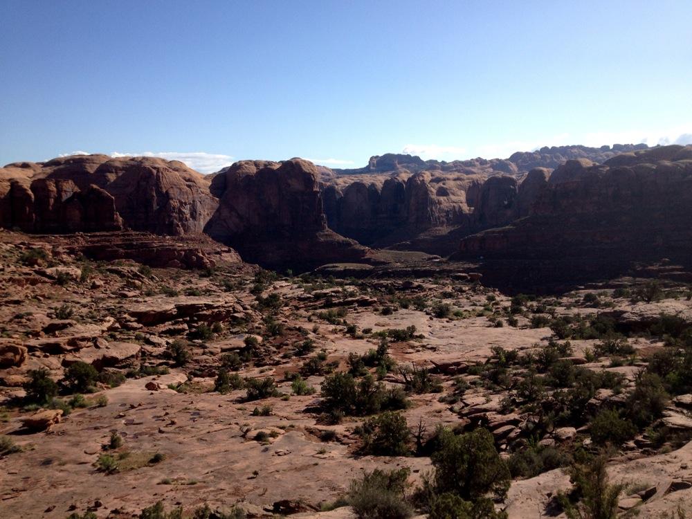 Expansive view of rugged red rock formations and desert landscape under a clear blue sky, with scattered vegetation and distant cliffs in the background. Hymasa mountain bike trail.