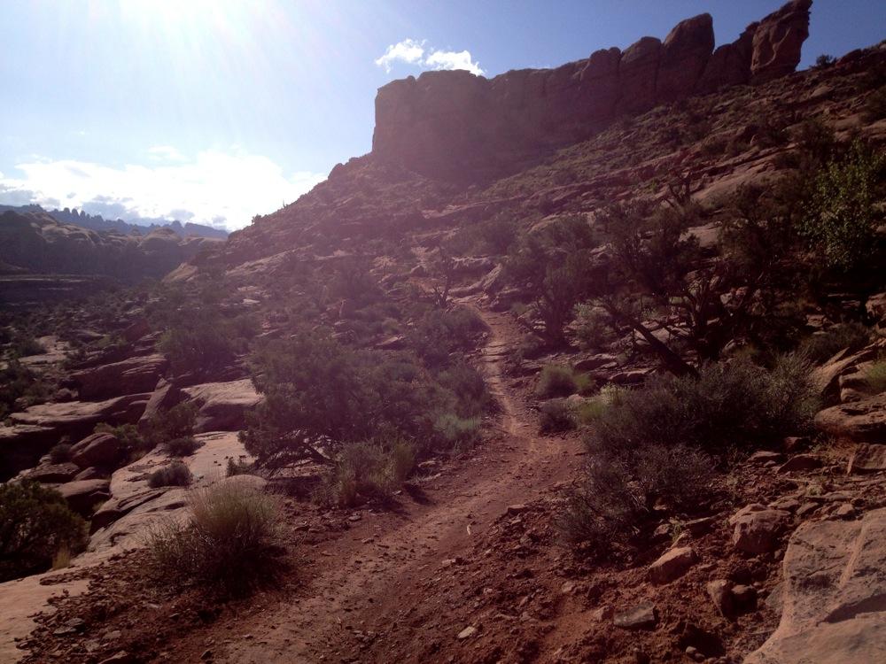 A winding dirt trail leads through a rocky landscape, surrounded by sparse vegetation and shrubs. The path gently ascends towards a prominent rock formation under a bright blue sky with scattered clouds. Sunlight casts a warm glow over the scene, highlighting the rugged terrain of the area. Hymasa mountain bike trail.