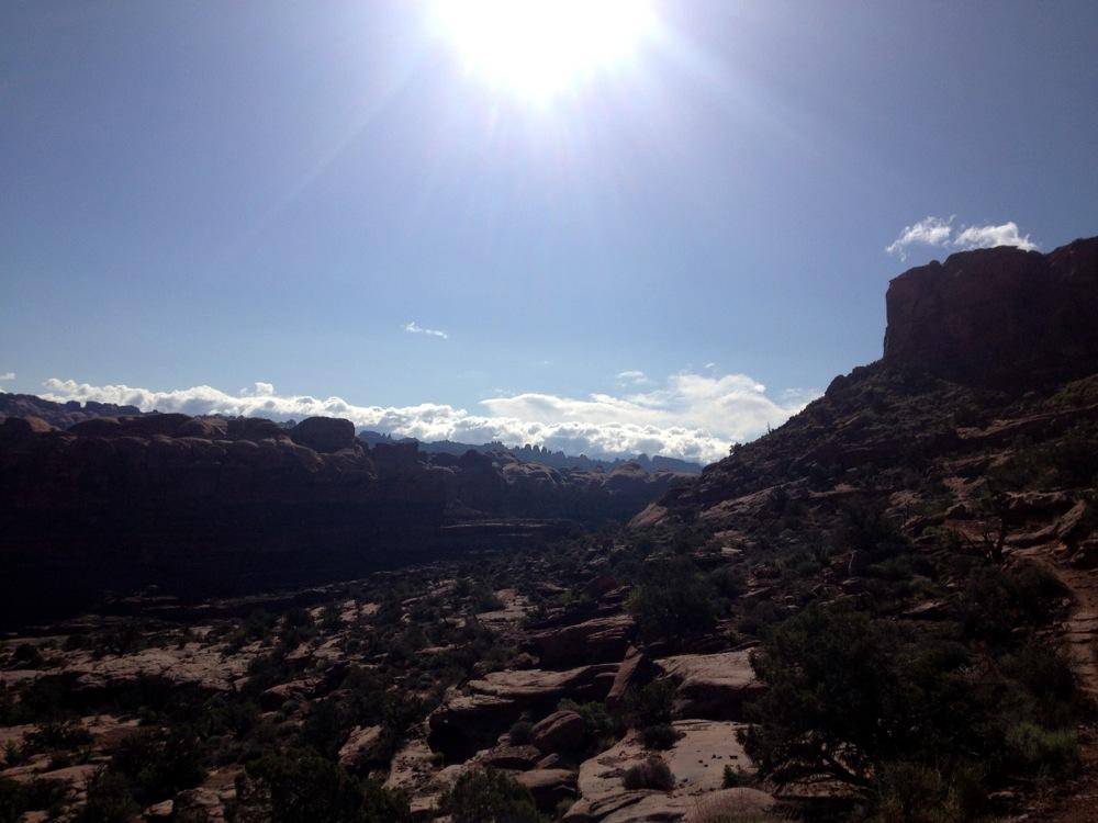 A sunlit landscape view showcasing rugged cliffs and rock formations under a bright sky, with scattered clouds in the distance. The foreground features sparse vegetation and rocky terrain, highlighting the natural beauty of the scenery. Hymasa mountain bike trail.