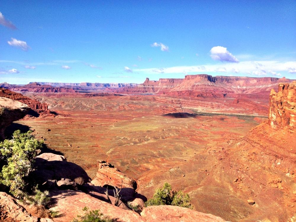 A vast landscape of red rock formations and mesas under a bright blue sky, showcasing the natural beauty of a desert environment. In the foreground, patches of green shrubs contrast with the arid terrain, while distant cliffs and dramatic geological features stretch across the horizon. Soft clouds dot the sky, adding depth to the scene. Hymasa mountain bike trail.