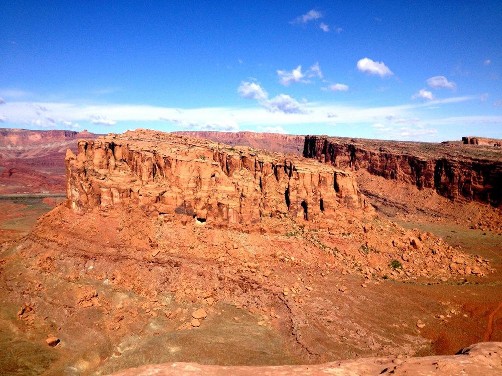 A panoramic view of a rugged desert landscape featuring a large sandstone butte surrounded by rocky terrain under a vibrant blue sky with scattered clouds. The warm reddish hues of the rocks contrast with the greenery in the valley below. Hymasa mountain bike trail.