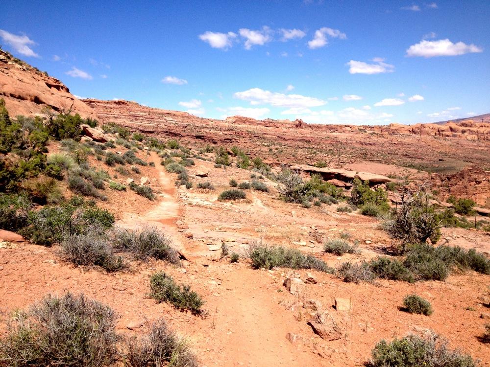 A scenic view of a rugged desert landscape featuring a winding dirt path leading through sparse vegetation. The terrain is characterized by reddish-brown rocky soil, scattered bushes, and distant rock formations under a bright blue sky with a few white clouds. Rockstacker mountain bike trail.