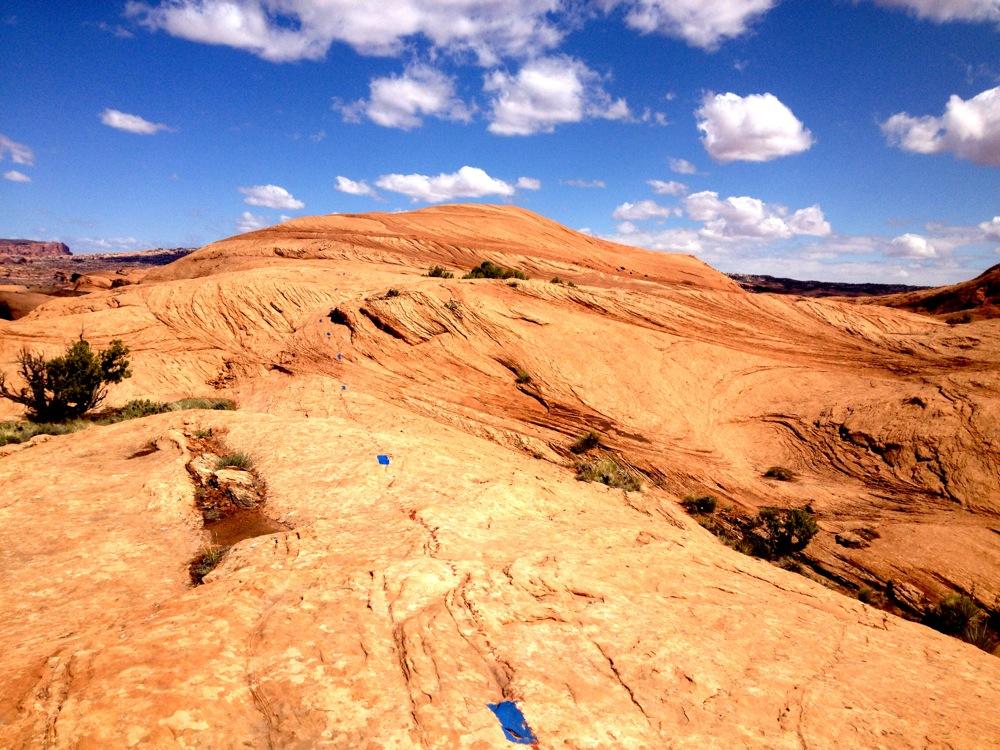 A panoramic view of a rocky desert landscape, featuring orange and tan sandstone formations with layered textures under a blue sky dotted with fluffy white clouds. Sparse greenery is visible along the rocky terrain, and blue markers are placed along the path. Pothole Arch Trail mountain bike trail.
