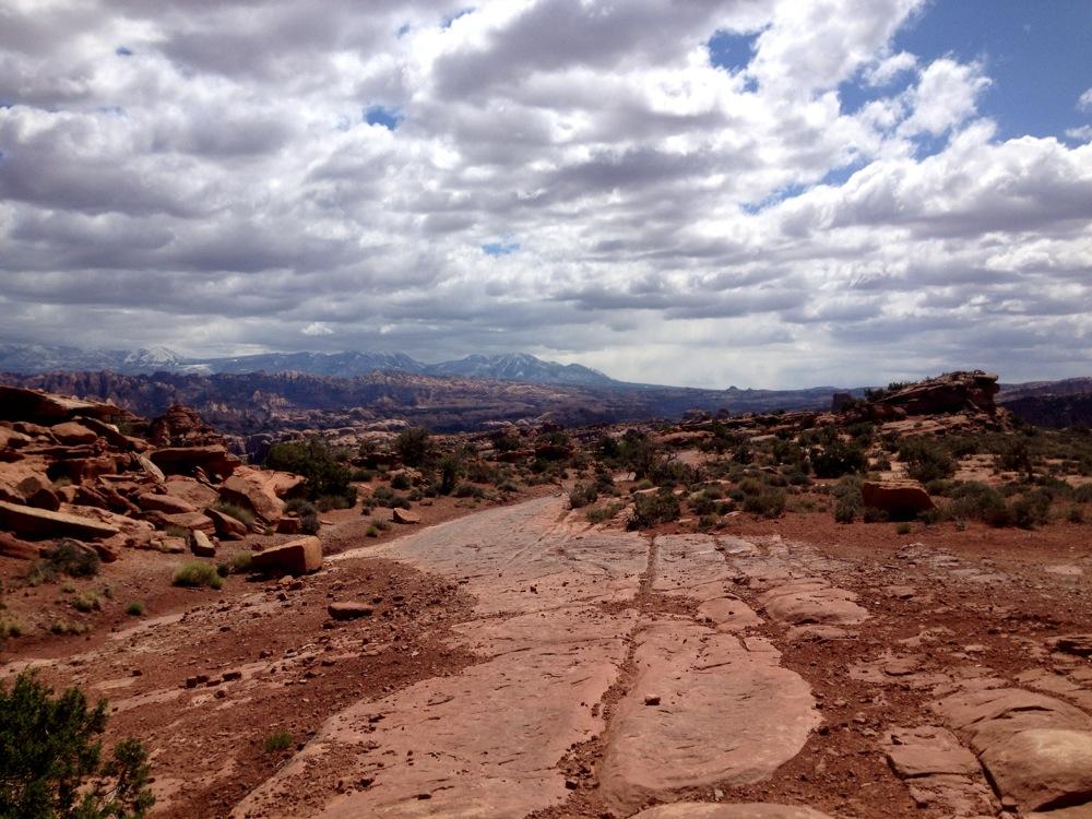 A rugged landscape featuring red rock formations and a dirt path, set against a backdrop of rolling hills and mountains under a cloudy sky. The terrain appears dry with sparse vegetation and rocky outcrops, suggesting a remote desert environment. Amasa Back Trail mountain bike trail.