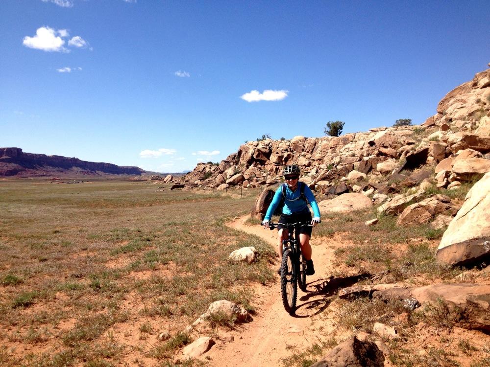 A person riding a mountain bike on a dirt trail surrounded by rocky terrain and open fields under a clear blue sky. Moab Brand Trails mountain bike trail.