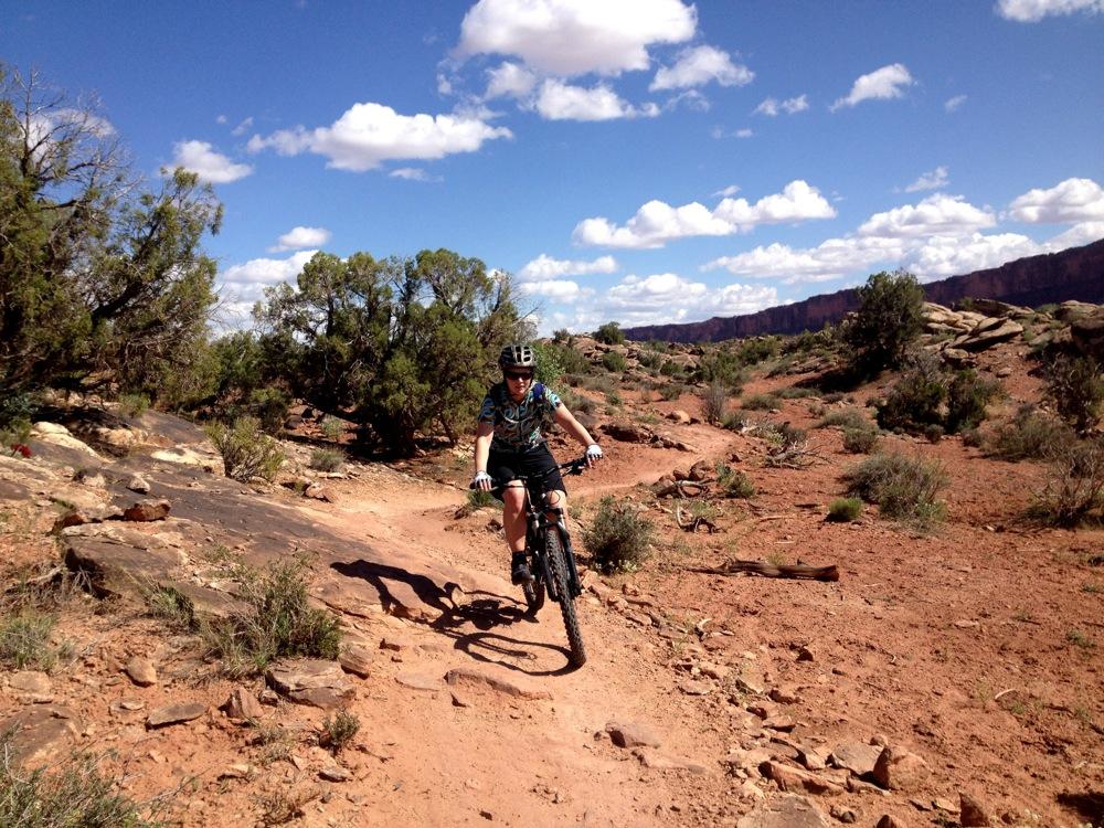 A person riding a mountain bike on a dirt trail surrounded by rocky terrain and sparse vegetation, under a partly cloudy blue sky. Moab Brand Trails mountain bike trail.