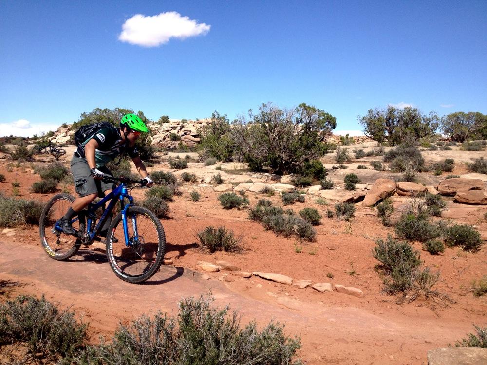 A mountain biker in a green helmet and cycling gear rides along a dirt trail surrounded by sparse vegetation and rocky terrain under a clear blue sky with a few clouds. Moab Brand Trails mountain bike trail.