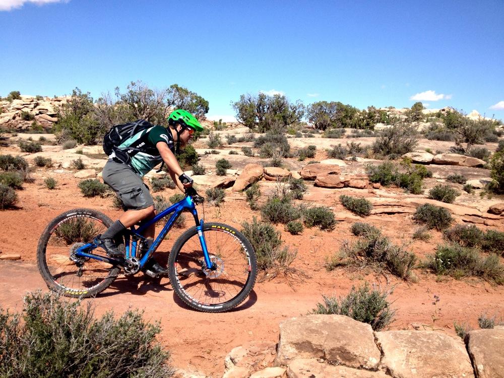 A person riding a blue mountain bike on a rocky trail in a desert landscape, wearing a green helmet and a backpack. The scene features dry vegetation and red sandy soil under a clear blue sky. Moab Brand Trails mountain bike trail.