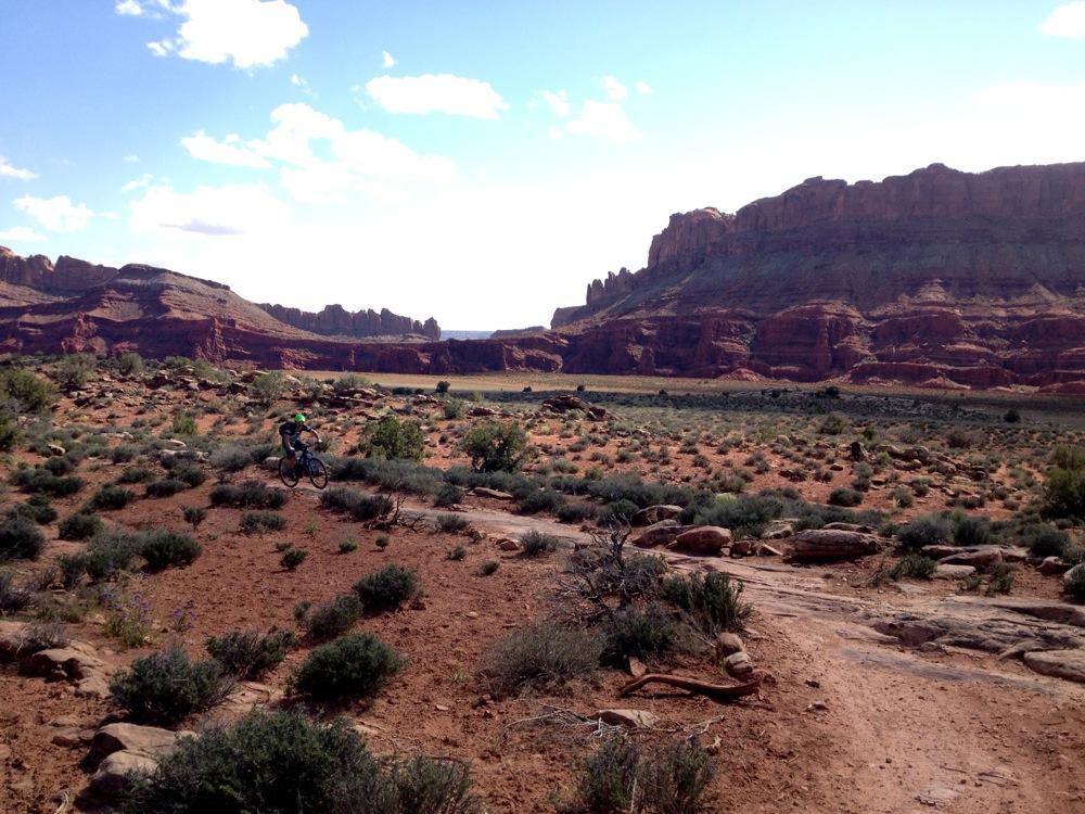A mountain biker rides along a dirt trail surrounded by rugged red rock formations and sparse vegetation under a clear blue sky with a few clouds. Moab Brand Trails mountain bike trail.