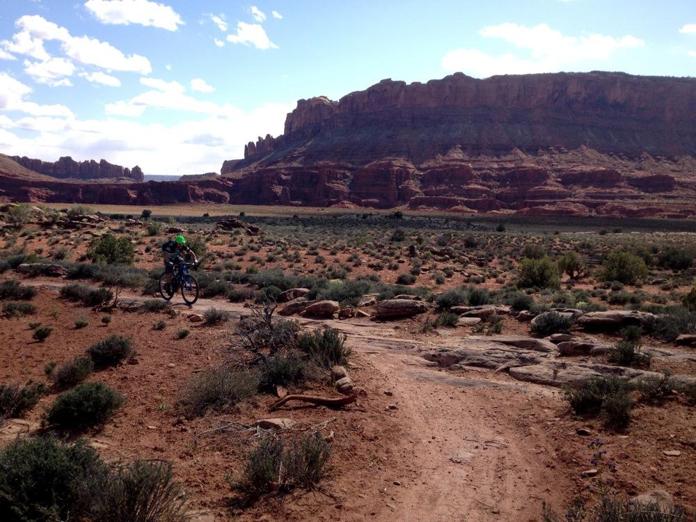 A person riding a mountain bike along a dirt trail in a rugged, arid landscape. The background features dramatic red rock formations and a blue sky with fluffy white clouds. Sparse vegetation, including shrubs and small bushes, is scattered across the terrain, highlighting the natural beauty of the desert environment. Moab Brand Trails mountain bike trail.