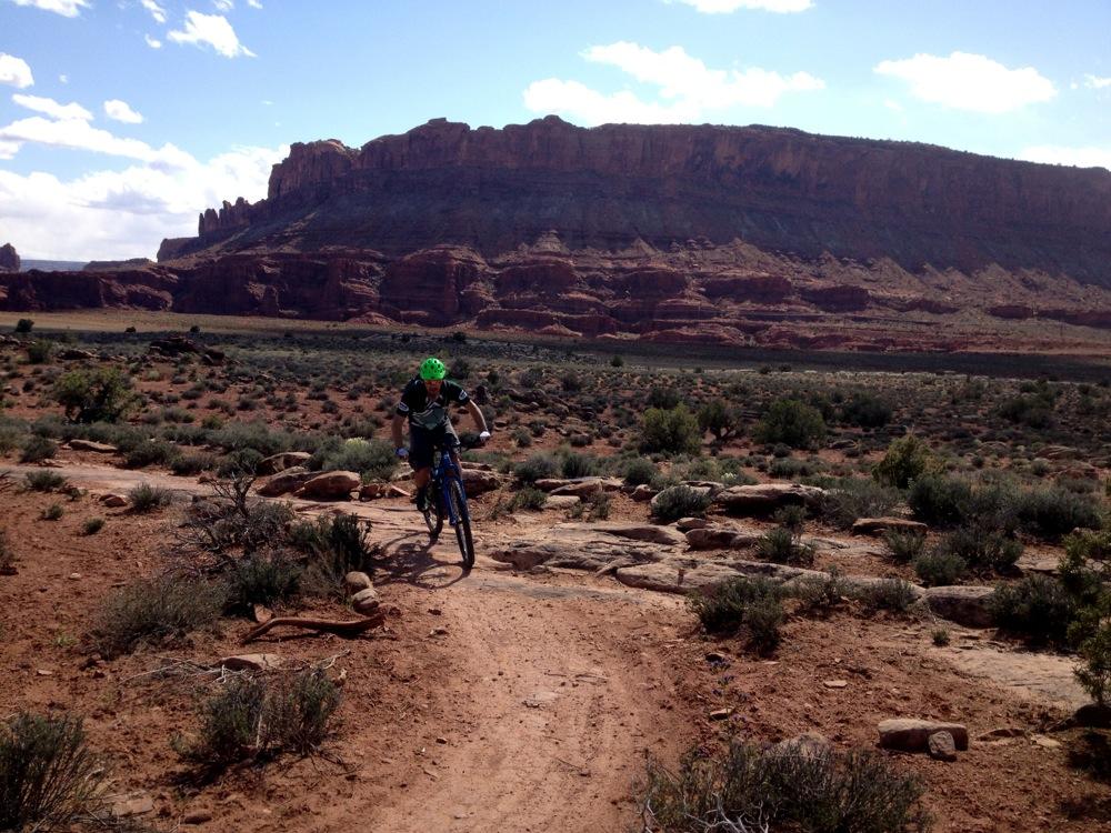 A person riding a mountain bike on a dirt trail surrounded by desert terrain, featuring rocky formations and shrubs, with steep cliffs in the background under a partly cloudy sky. Moab Brand Trails mountain bike trail.