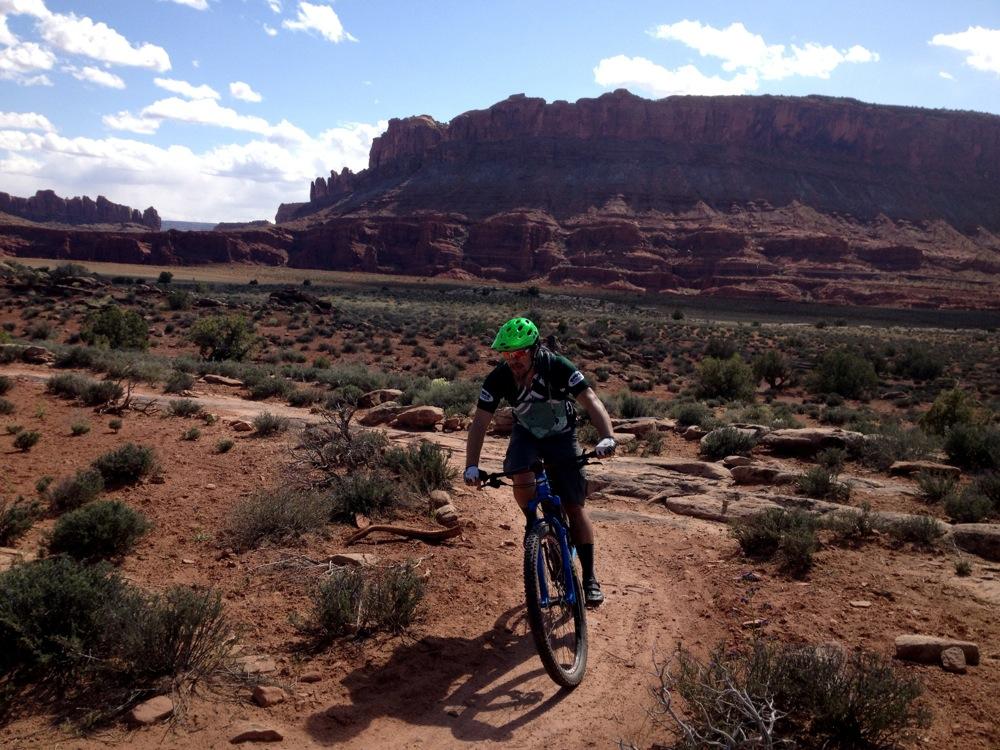 A cyclist riding a mountain bike on a rugged dirt trail surrounded by desert landscape, featuring red rock formations and sparse vegetation under a partly cloudy sky. Moab Brand Trails mountain bike trail.