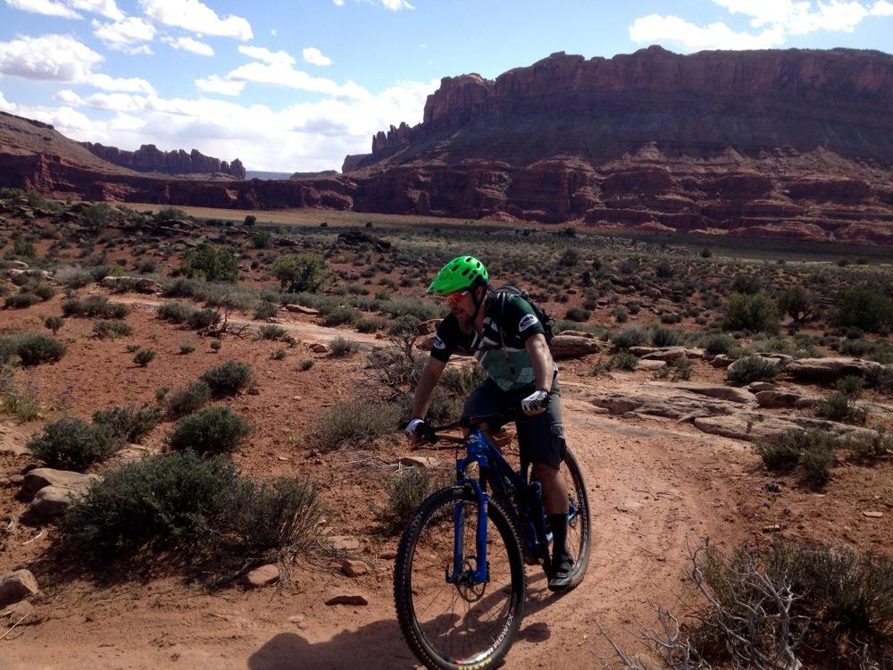 A mountain biker in a green helmet navigates a dirt trail surrounded by desert landscape, featuring red rock formations and scattered vegetation under a partly cloudy sky. Moab Brand Trails mountain bike trail.