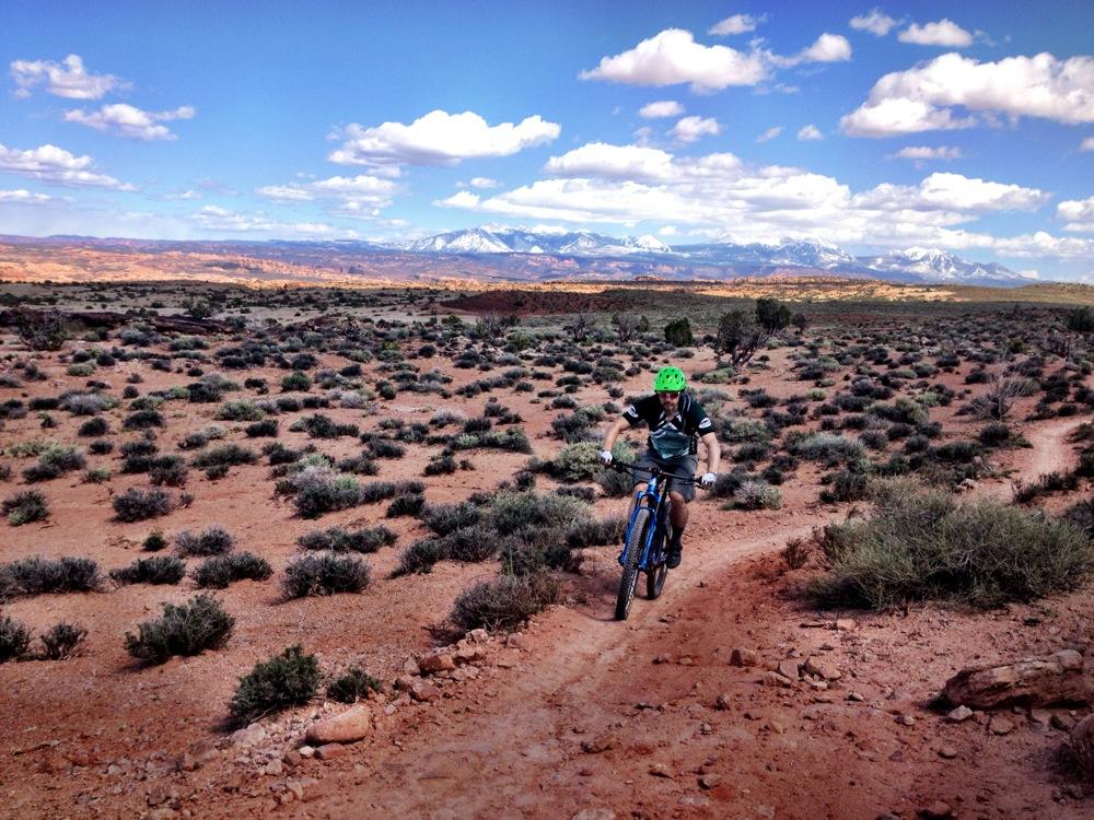 A person riding a mountain bike on a dirt trail surrounded by sparse vegetation and rocky terrain, with a backdrop of distant mountains and a blue sky filled with fluffy white clouds. Moab Brand Trails mountain bike trail.