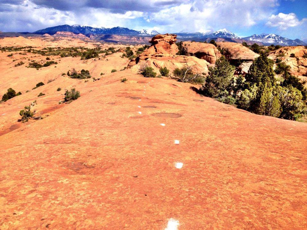 A scenic landscape featuring a rocky terrain with reddish sandstone, dotted with patches of vegetation. In the background, mountains are visible under a partly cloudy sky. White markings can be seen along the rock surface, indicating a possible trail. Slickrock mountain bike trail.