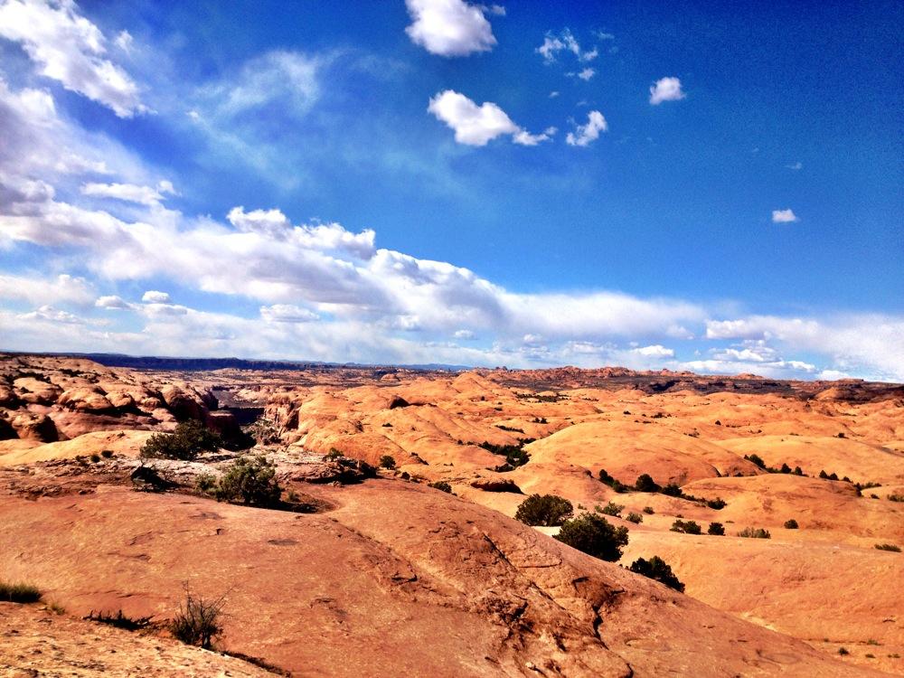 A panoramic view of a desert landscape featuring sandy hills and rocky formations under a blue sky with scattered clouds. Small patches of greenery are visible amidst the rugged terrain. Slickrock mountain bike trail.