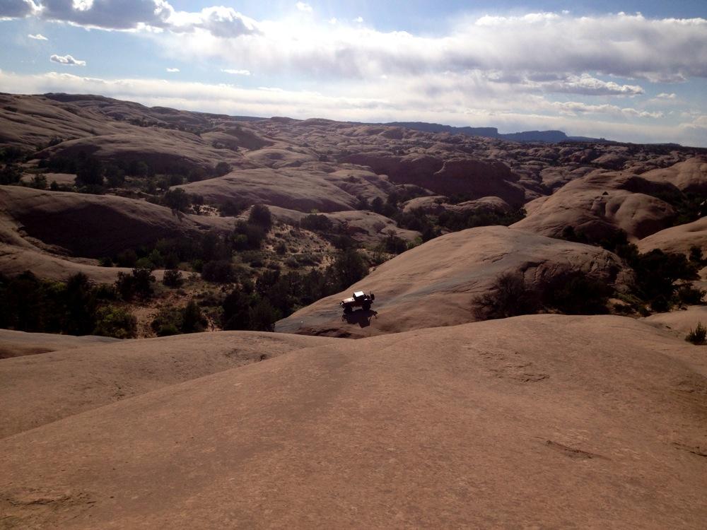 A rugged landscape featuring smooth, hilly terrain with a vehicle parked on a rocky surface. The scene is illuminated by sunlight, with distant mountains visible under a partly cloudy sky. Green shrubs and trees are scattered throughout the area. Fins n Things mountain bike trail.