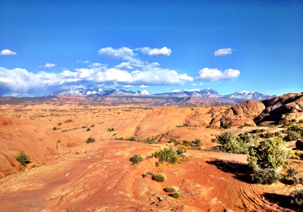 A panoramic view of a rugged desert landscape featuring red rock formations and sparse vegetation, with snow-capped mountains in the background under a bright blue sky dotted with white clouds. Slickrock mountain bike trail.