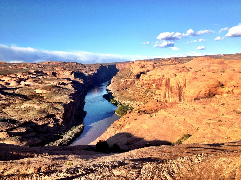 A panoramic view of a winding river surrounded by rugged red rock formations under a clear blue sky, with a mix of green vegetation visible along the riverbanks. The landscape features steep cliffs and a varied terrain, showcasing the natural beauty of a canyon setting. Slickrock mountain bike trail.