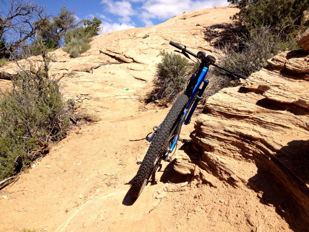 A mountain bike positioned on a rocky, sloped path surrounded by sparse vegetation under a bright blue sky. Rocky Tops mountain bike trail.