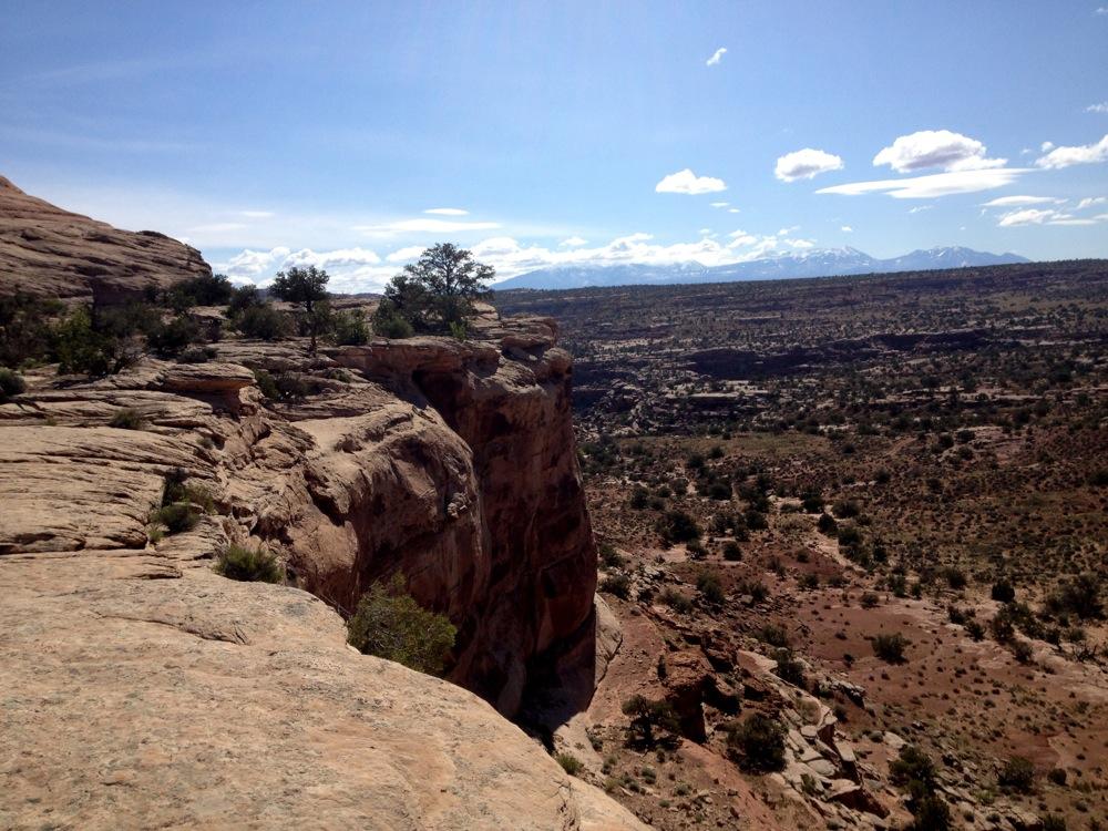 A scenic view of a rocky landscape, featuring a cliff edge with sparse vegetation and a distant mountain range under a bright blue sky with scattered clouds. The textured rock formations and desert terrain are visible, emphasizing the rugged beauty of the natural environment. Rocky Tops mountain bike trail.