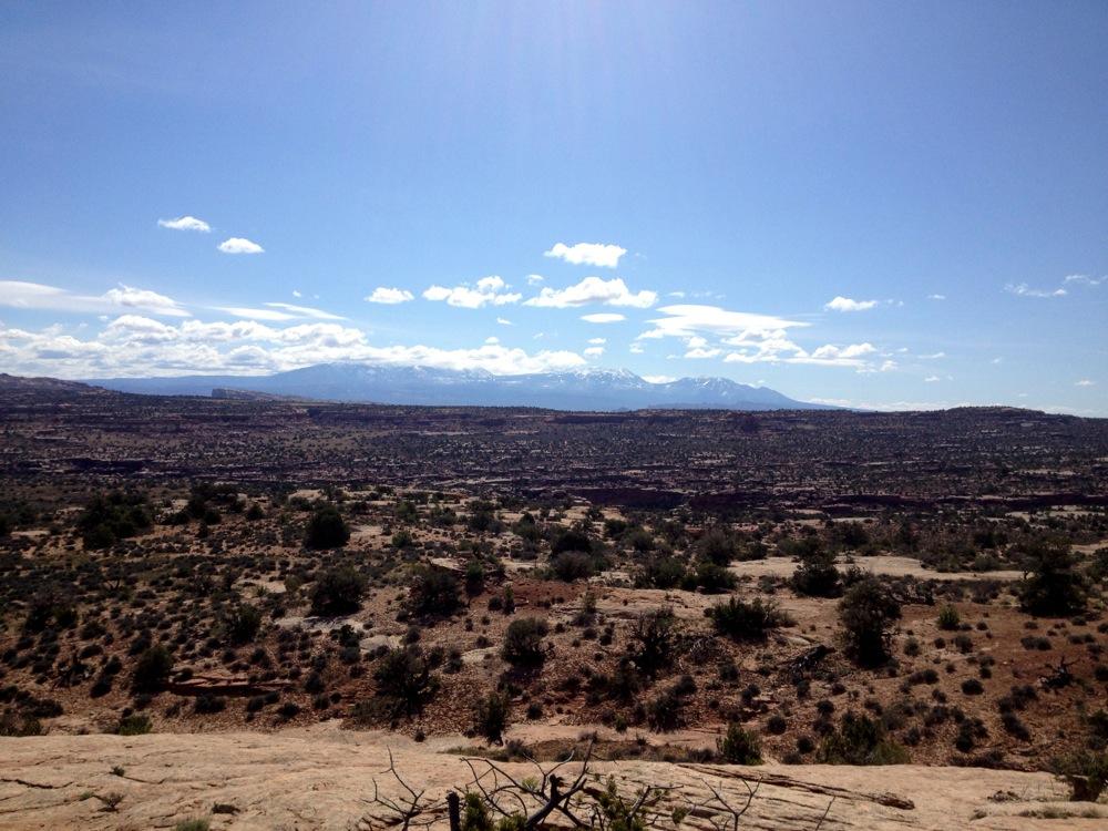 A panoramic view of a desert landscape featuring rock formations, sparse shrubs, and distant snow-capped mountains under a clear blue sky with scattered clouds. Rocky Tops mountain bike trail.