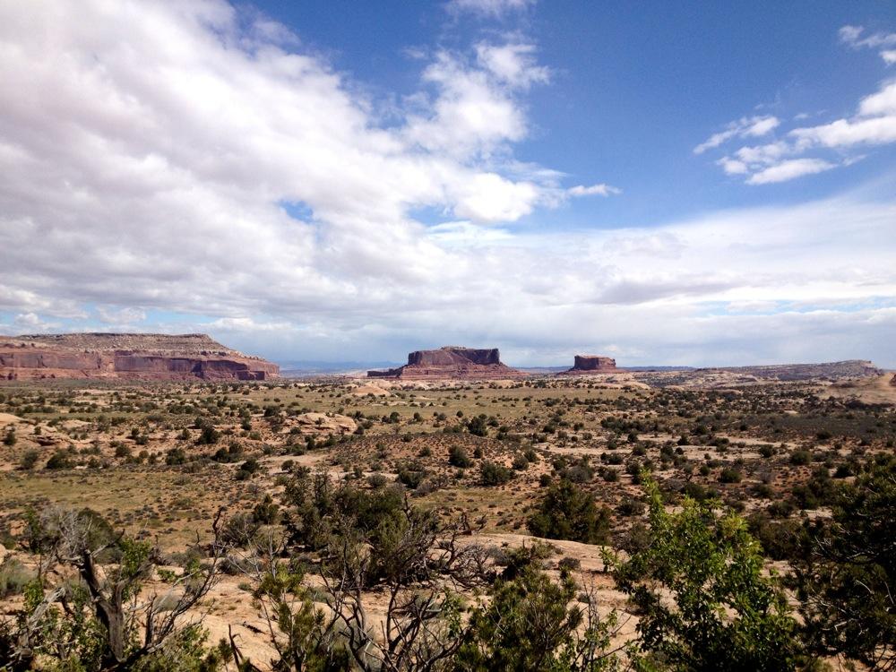 A panoramic view of a vast desert landscape featuring rugged mesas and rocky cliffs, under a partly cloudy sky. Sparse vegetation dots the foreground, adding to the arid, natural scenery. Rocky Tops mountain bike trail.