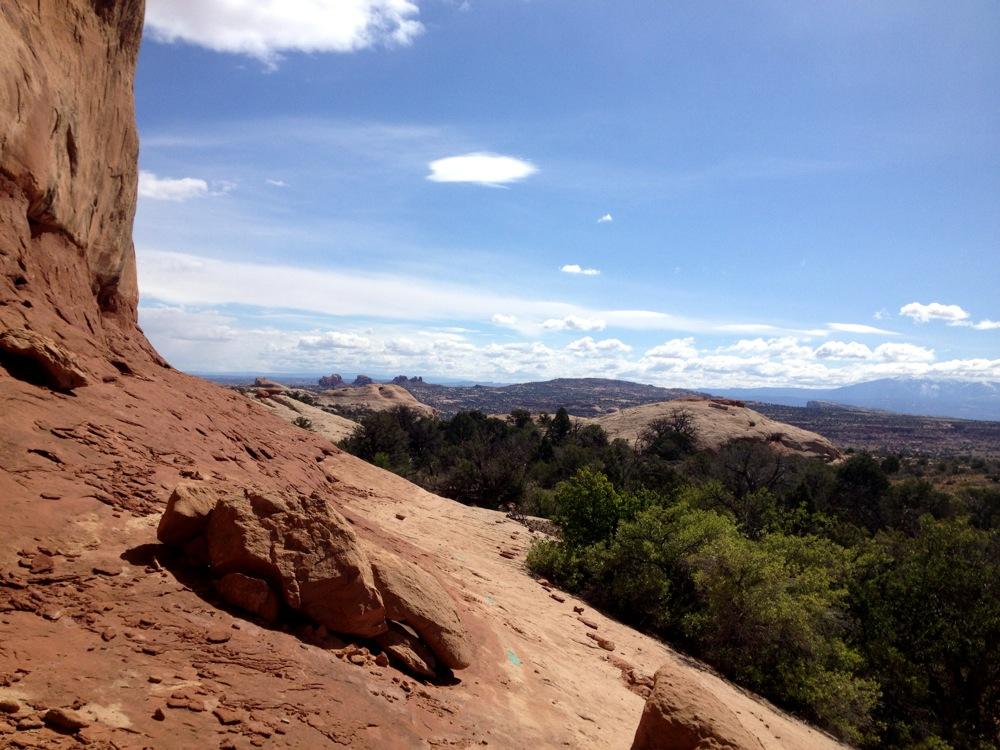 A scenic view of red rock formations and distant hills under a blue sky with scattered clouds. The foreground features rocky terrain and patches of green vegetation. Rocky Tops mountain bike trail.
