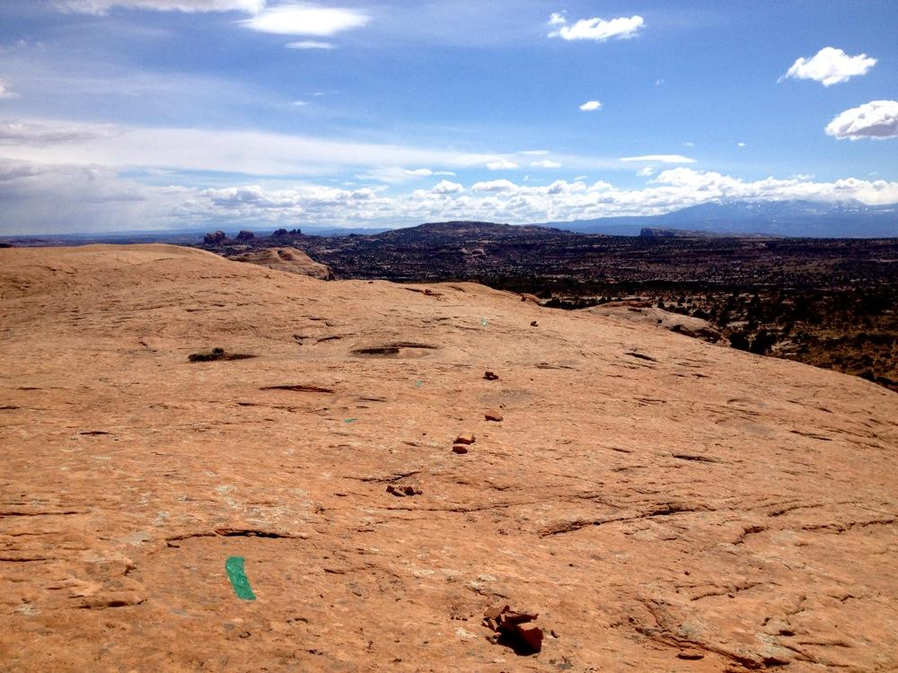A panoramic view of a rocky desert landscape under a partly cloudy sky, showcasing a flat, reddish-brown stone surface with scattered vegetation and markings on the rock. In the distance, rolling hills and rocky formations can be seen, extending towards a blue horizon. Rocky Tops mountain bike trail.