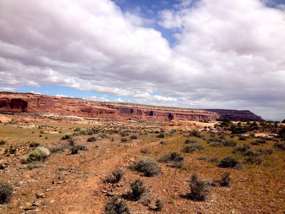 A panoramic view of a rugged desert landscape featuring reddish rock formations under a partly cloudy sky. The foreground includes sparse vegetation and a dirt path, leading towards the distant cliffs that rise dramatically against the horizon. Rocky Tops mountain bike trail.