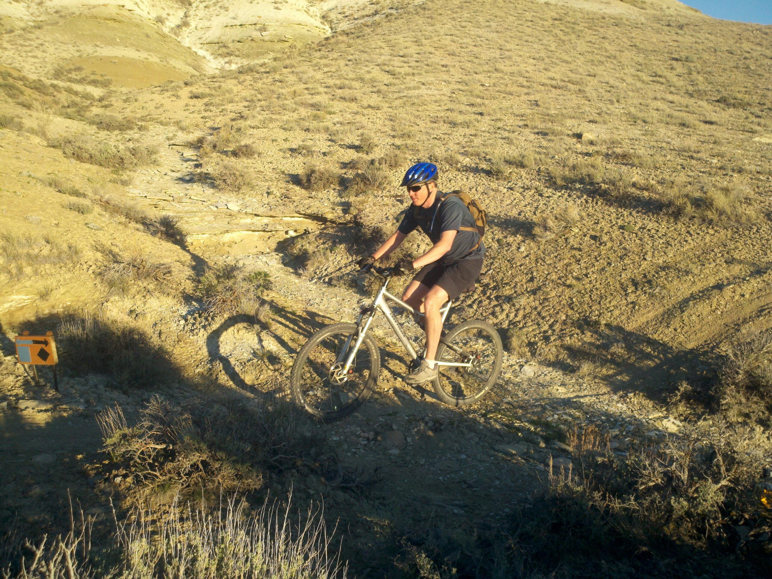 A mountain biker navigating a rocky trail in a dry, hilly landscape. The cyclist is wearing a blue helmet and a backpack, focusing on the path ahead. An orange directional sign is visible nearby, indicating the route. Pansy mountain bike trail.
